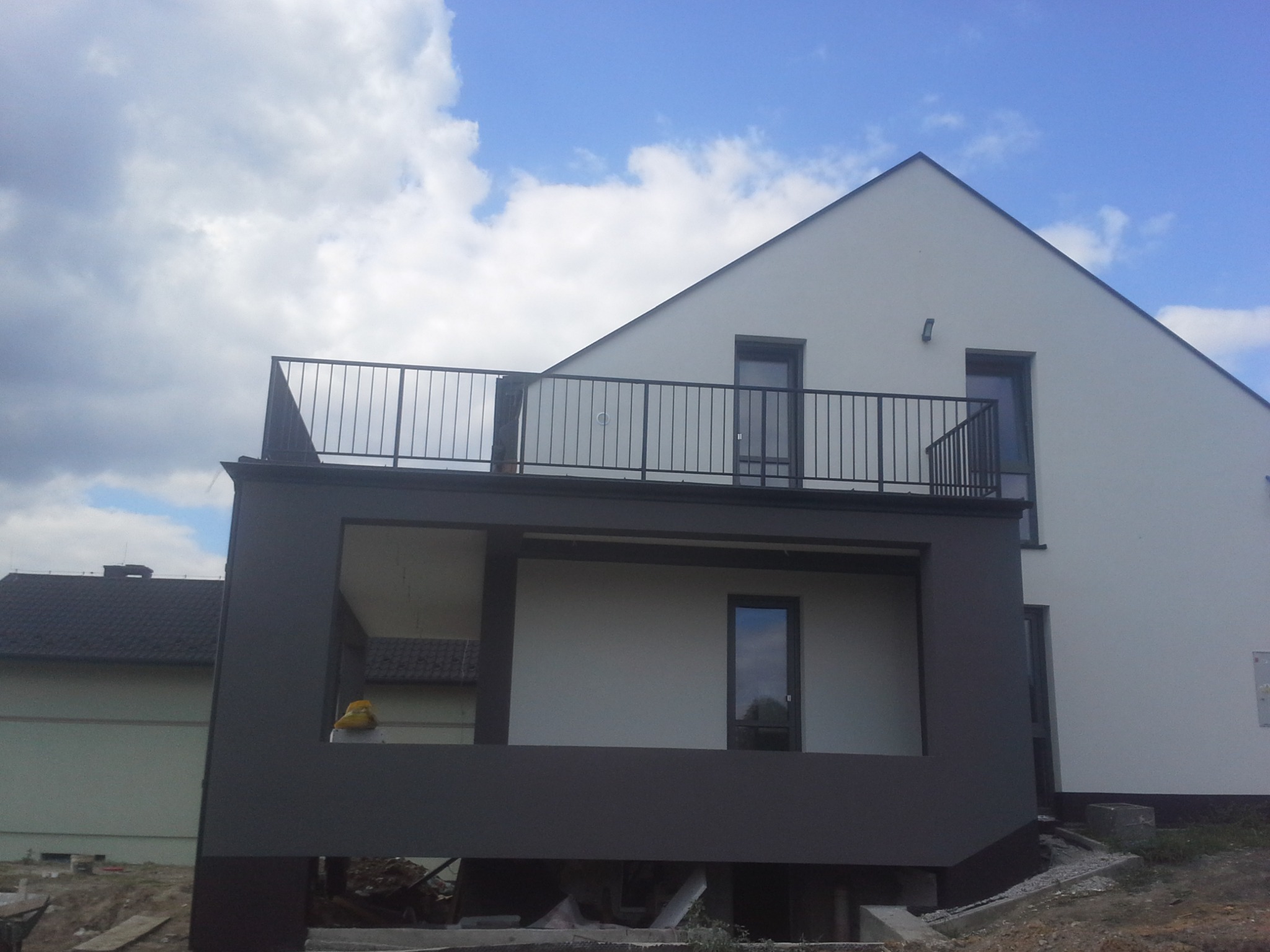 Metalowa balustrada na balkonie nowoczesnego domu z widokiem na błękitne niebo z chmurami, widoczne elementy konstrukcyjne budynku w trakcie budowy.