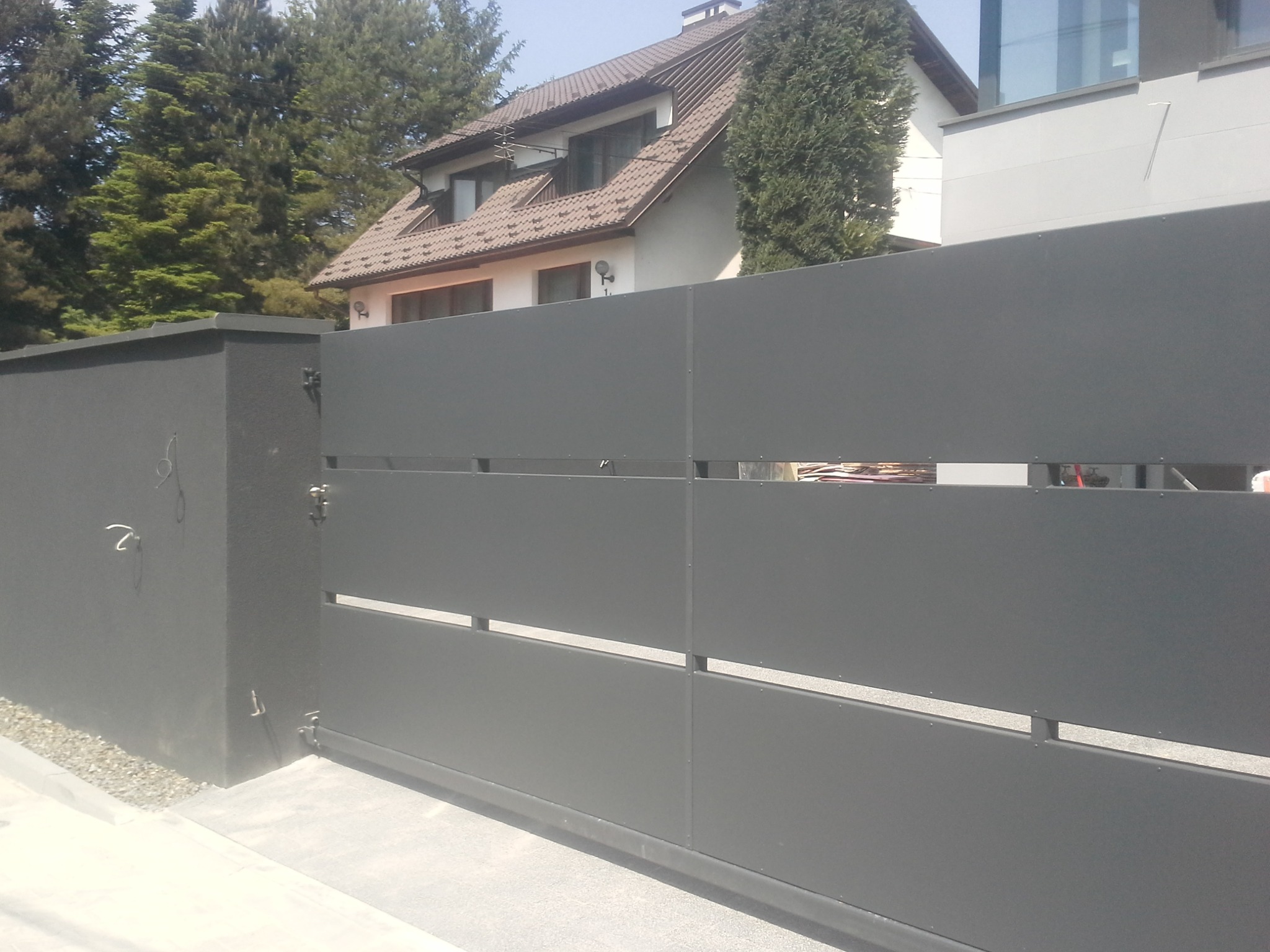 Nowoczesna, przesuwna brama w kolorze grafitowym z poziomymi prześwitami, zamontowana przed domem z brązowym dachem i jasną elewacją.