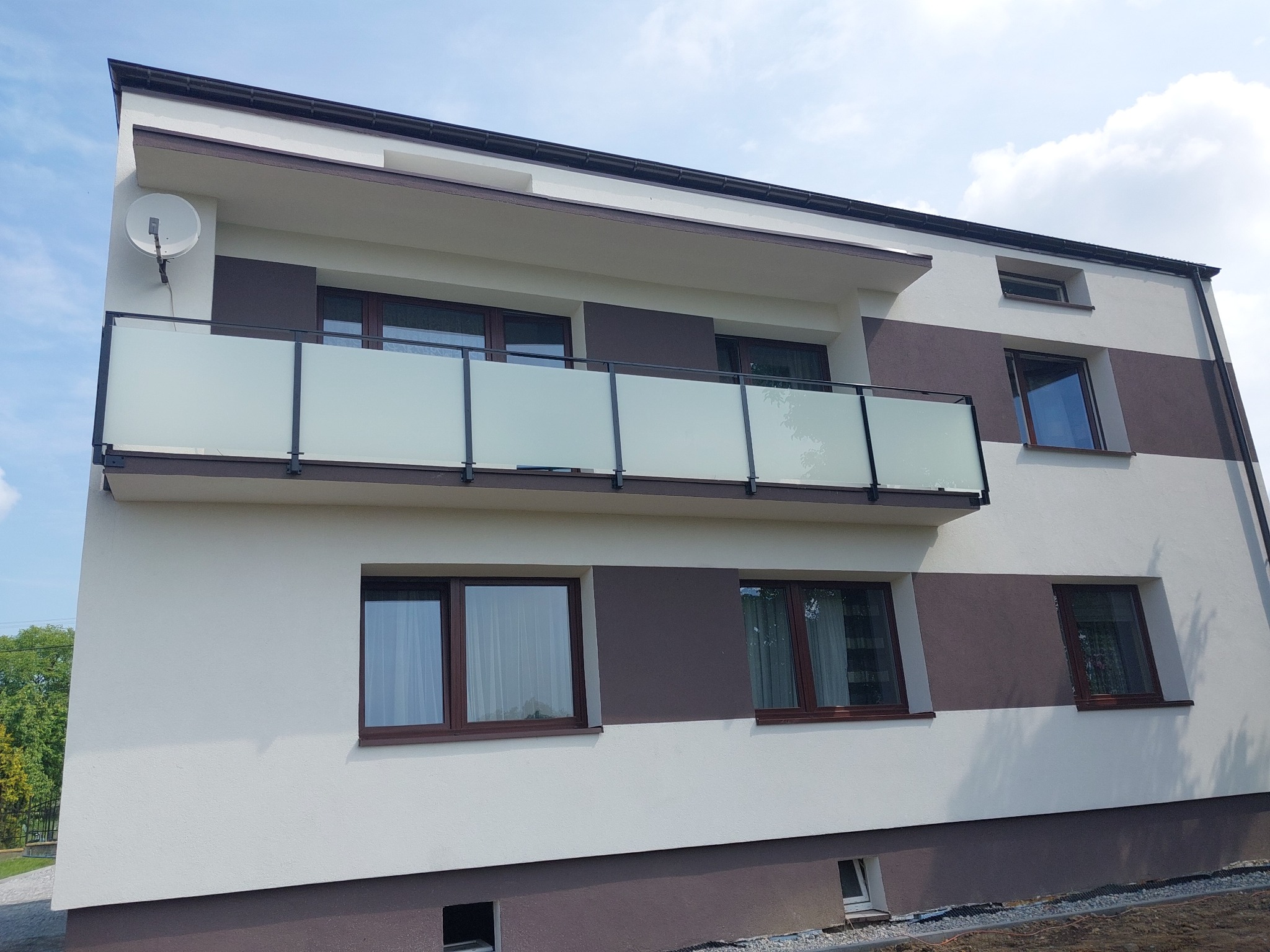 Nowoczesny budynek z balkonem zabezpieczonym mleczną szklaną balustradą z czarnymi metalowymi ramami, na elewacji widoczna antena satelitarna.