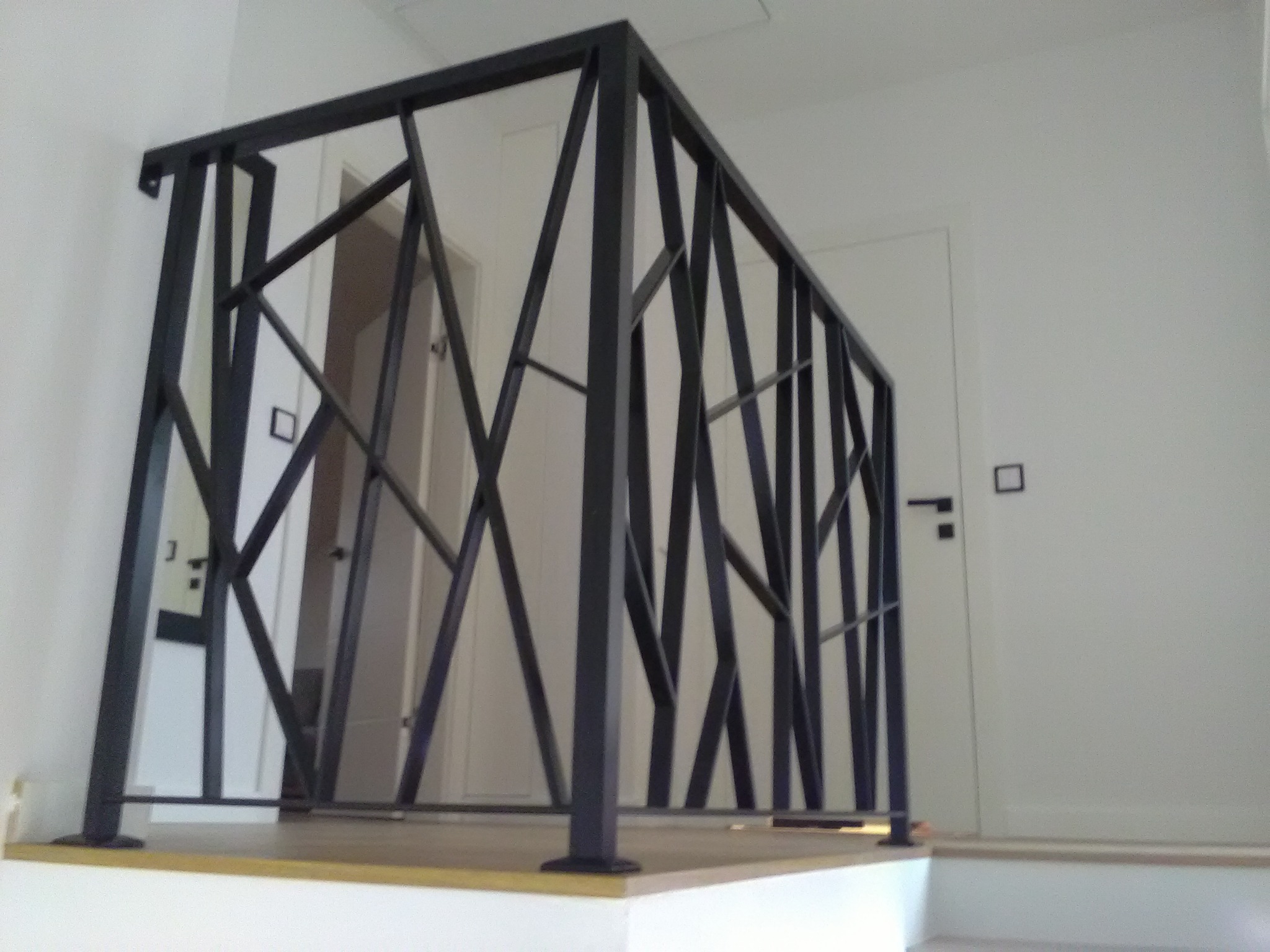 Czarna balustrada o geometrycznym wzorze, z ukośnymi i pionowymi prętami, zamontowana na antresoli w nowoczesnym wnętrzu z białymi ścianami i drzwiami.