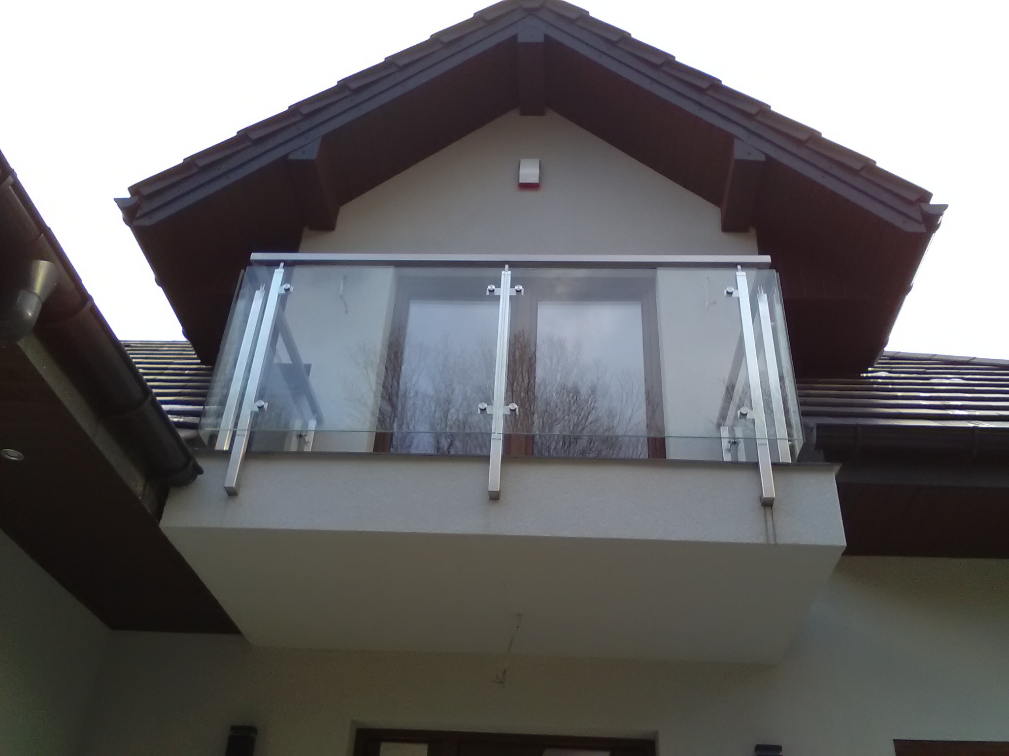 Zewnętrzny balkon z nowoczesną szklaną balustradą, mocowaną na metalowych słupkach, widoczny z dołu na tle dwuspadowego dachu budynku.