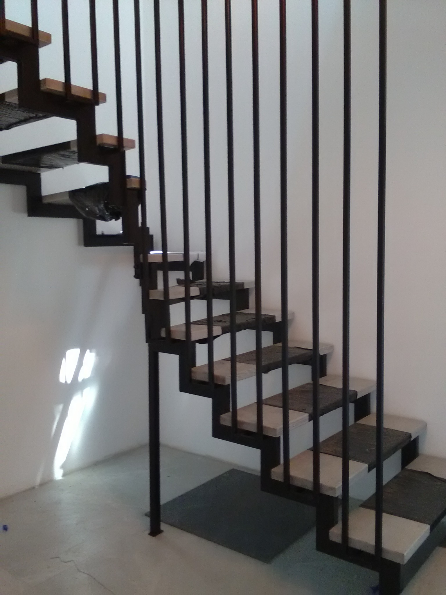 Nowoczesne schody metalowe z czarną konstrukcją i jasnymi stopniami, zabezpieczone pionowymi prętami, widoczne w surowym wnętrzu z białymi ścianami i betonową podłogą.