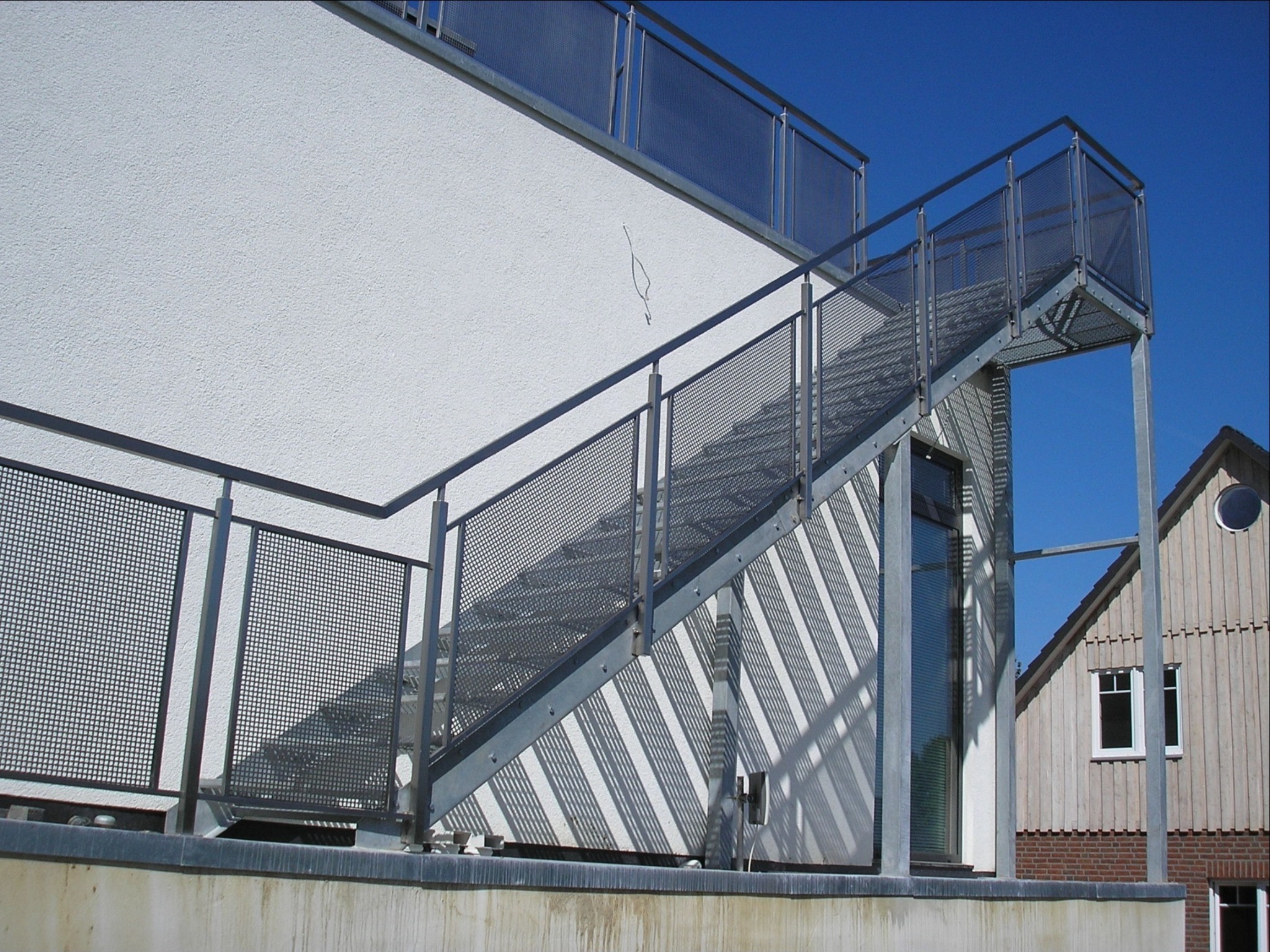 Zewnętrzne schody ewakuacyjne z metalową siatką i poręczą, przymocowane do budynku z białą elewacją, widok z dołu na tle błękitnego nieba i budynku obok.