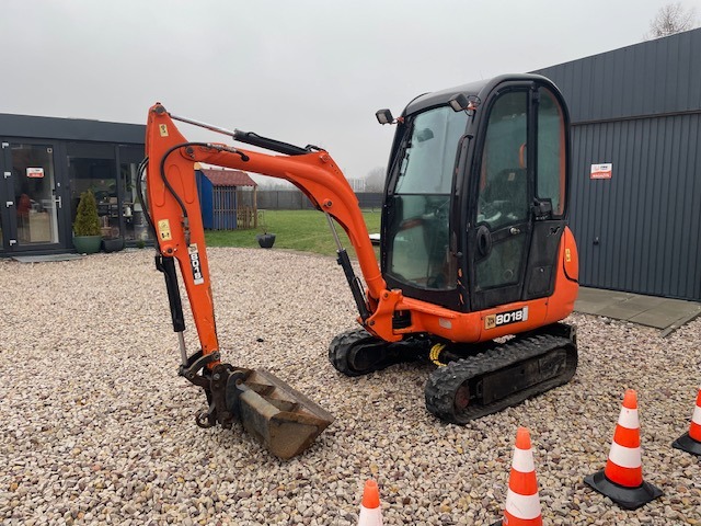 Pomarańczowa mini koparka JCB 8018 na żwirowym podłożu, z widocznymi pachołkami ostrzegawczymi, budynkiem w tle i pochmurnym niebem.