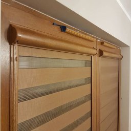 Janusz Matera - Zamknięte rolety dzień noc w drewnianej ramie okiennej, z widocznym mechanizmem sterującym. Beżowo-brązowe pasy tworzą efekt zaciemnienia.