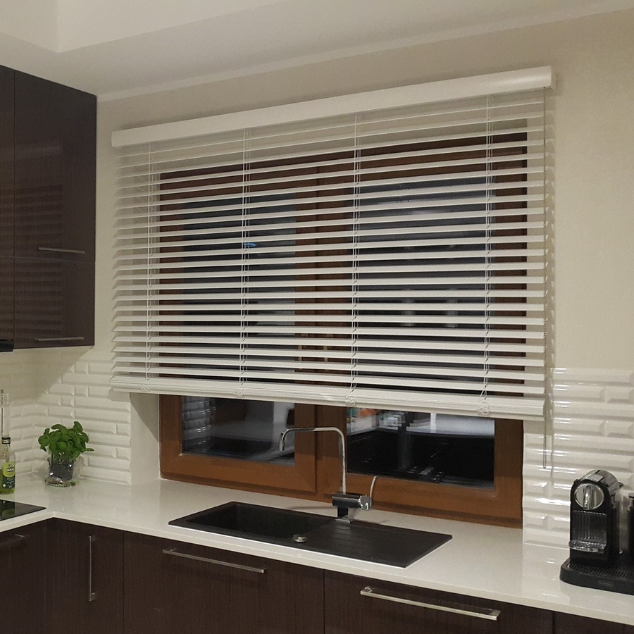 Kuchnia z białymi żaluzjami poziomymi w oknie o drewnianej ramie, nad zlewem. Widoczne elementy wyposażenia kuchni i dekoracyjna ściana.