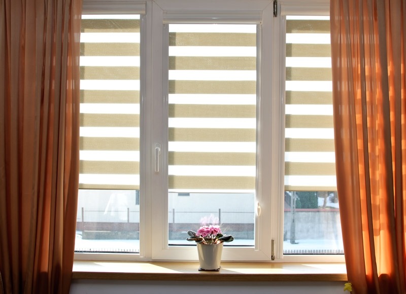 Beżowe rolety dzień noc zamontowane w białym oknie trzyskrzydłowym, z zasłonami po bokach. Na parapecie doniczka z fioletowymi kwiatami.