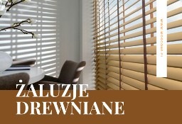 Beżowe żaluzje drewniane z ciemnobrązowymi taśmami, zamontowane w jasnym wnętrzu. Na parapecie dekoracja z gałązek kwitnącej rośliny.