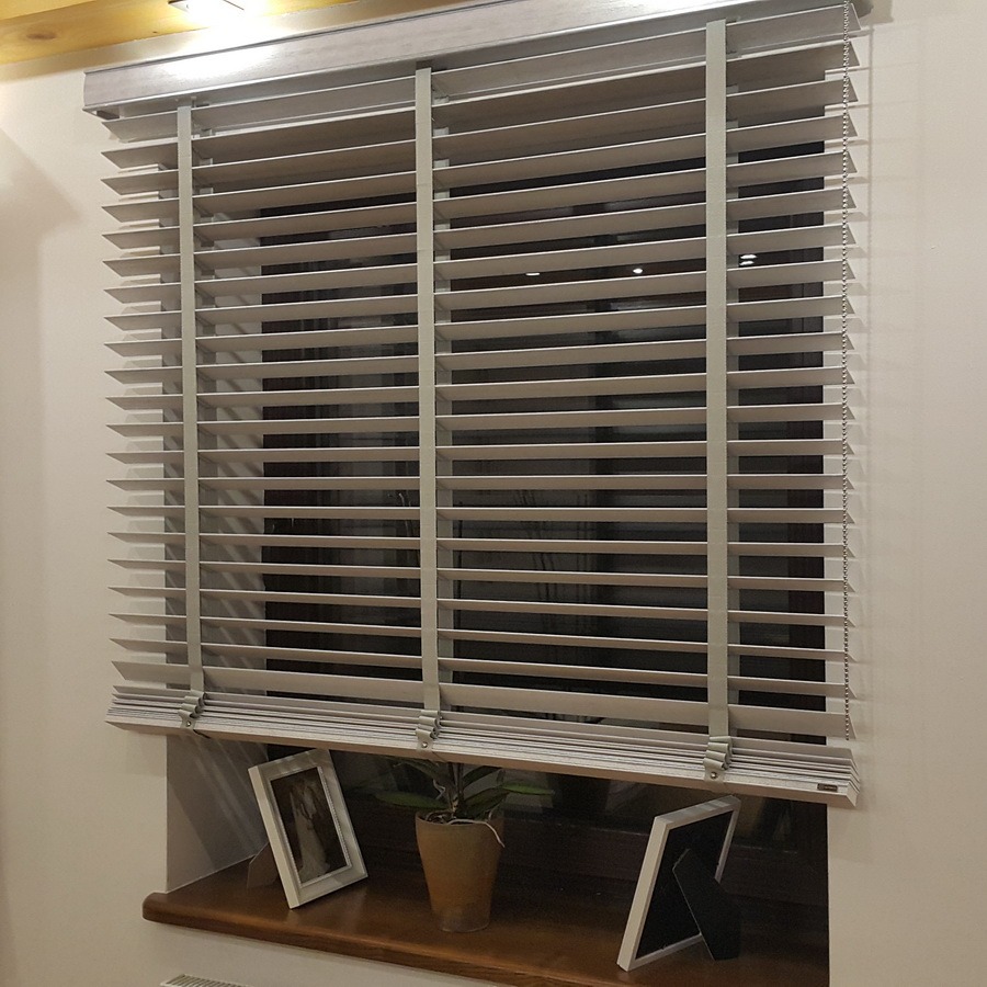 Zamknięte, poziome żaluzje drewniane w odcieniach szarości zamontowane w oknie. Widoczne ramki ze zdjęciami i roślina doniczkowa na parapecie.