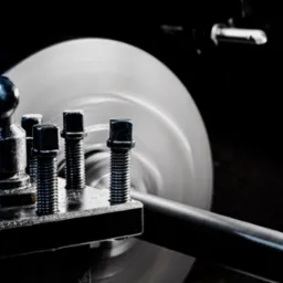 Metalowy pręt obrabiany na tokarce, widoczne śruby mocujące i wirujący element obrabiany w tle, ciemne otoczenie.