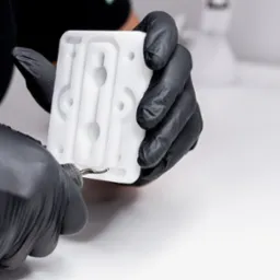 Operator w czarnych rękawicach precyzyjnie obrabia frezem krawędź białego, plastikowego elementu z licznymi otworami na białym tle.