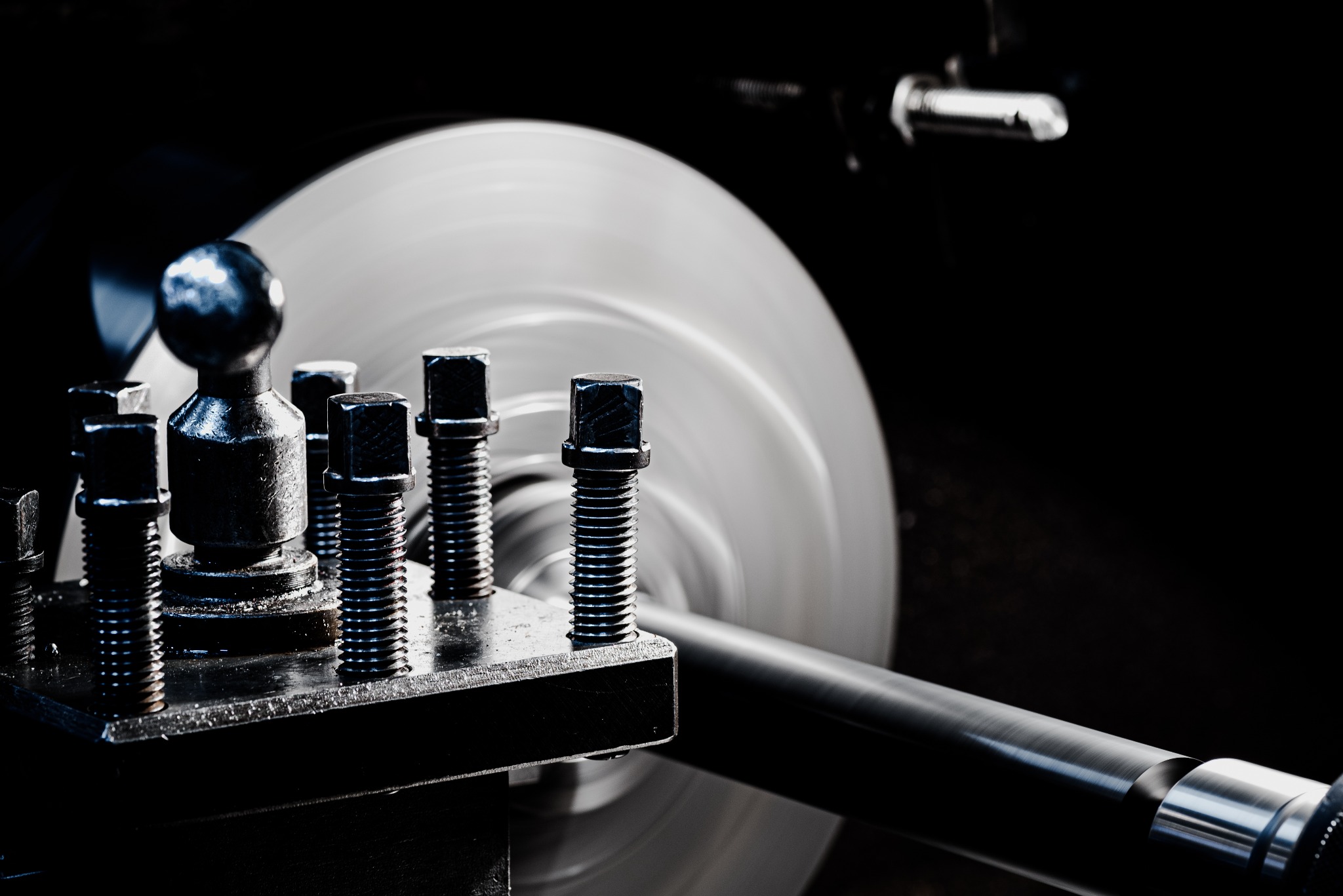 Metalowy pręt obrabiany na tokarce, widoczne śruby mocujące i wirujący element obrabiany w tle, ciemne otoczenie.