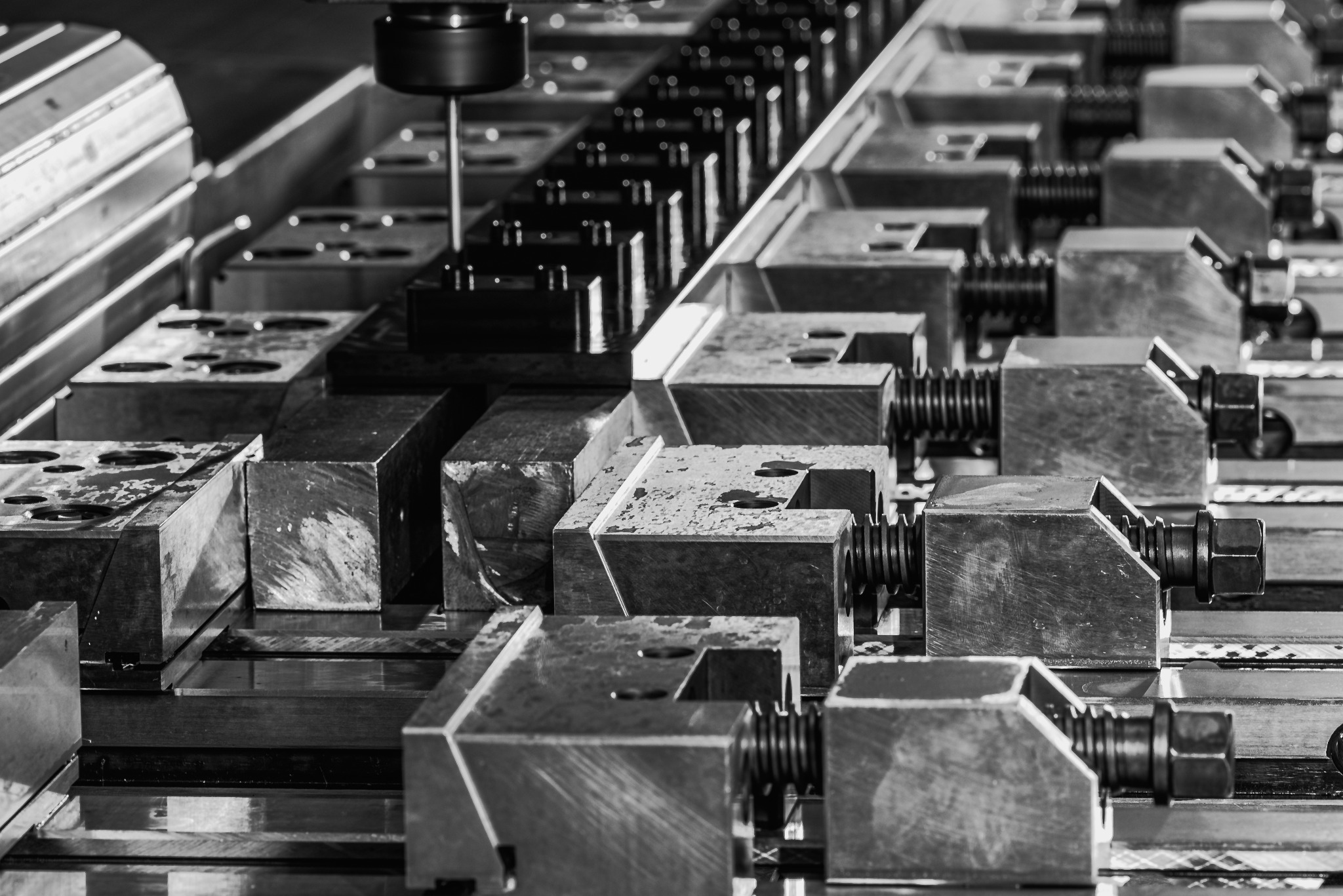 Czarno-białe zdjęcie detalu frezarki CNC z zamocowanymi w uchwytach surowymi, metalowymi blokami przed obróbką, widoczne ślady chłodziwa.