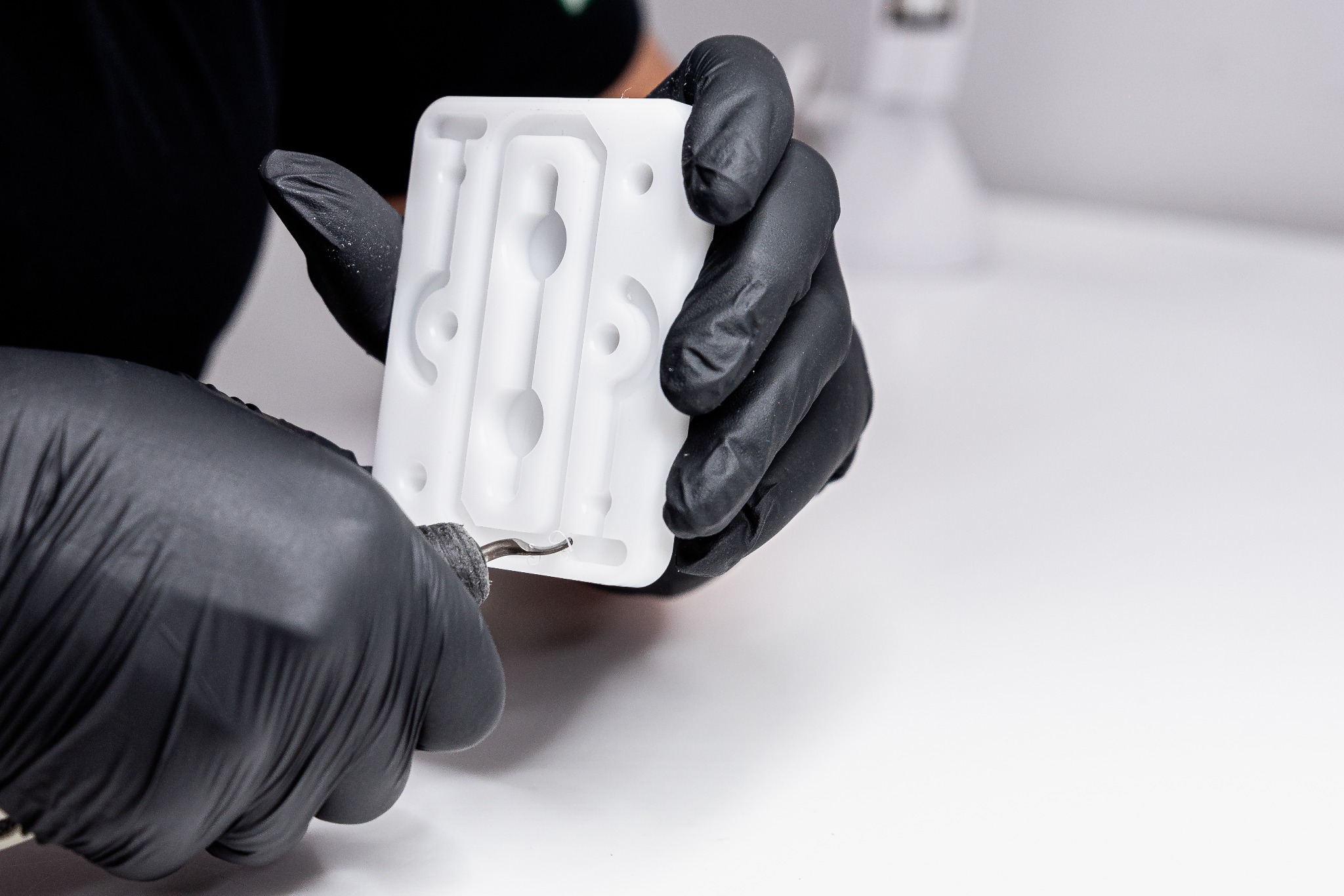 Operator w czarnych rękawicach precyzyjnie obrabia frezem krawędź białego, plastikowego elementu z licznymi otworami na białym tle.