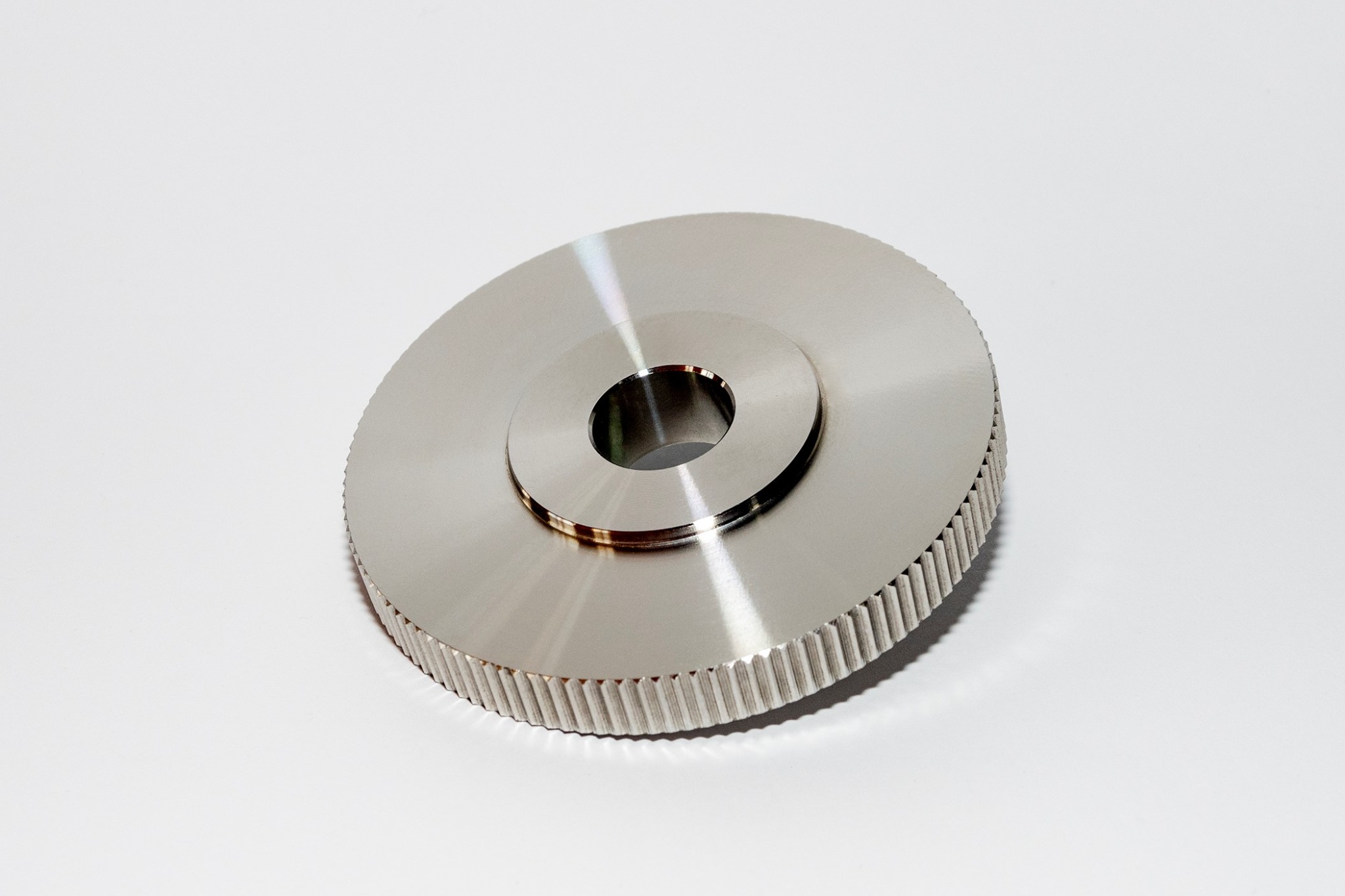 Metalowy element z centralnym otworem i radełkowanym obwodem, leżący na białym tle pod delikatnym kątem.