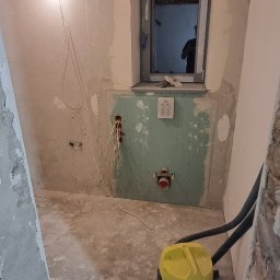 MEL- FI Remonty - Łazienka w trakcie remontu, widoczne ściany przygotowane do położenia płytek, wystające kable, okno z widocznym odbiciem osoby oraz żółty odkurzacz przemysłowy.