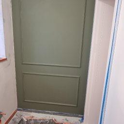 MEL- FI Remonty - Ściana pomalowana na oliwkowy kolor z dekoracyjnymi panelami, zabezpieczona folią malarską na podłodze, widoczna dziura w suficie i taśma malarska na framudze drzwi.