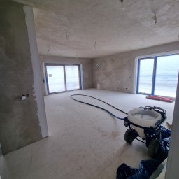 MEL- FI Remonty - Wnętrze w trakcie prac przygotowawczych do wykończenia, ściany i sufit pokryte świeżą warstwą gładzi, okna zabezpieczone folią, na podłodze maszyna do nakładania gładzi z niebieskim wężem.