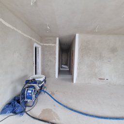 MEL- FI Remonty - Wnętrze w trakcie prac wykończeniowych, widoczny agregat malarski T-Max 506 na tle świeżo nałożonych gładzi na ścianach i suficie, z widocznymi przewodami elektrycznymi wystającymi z sufitu.