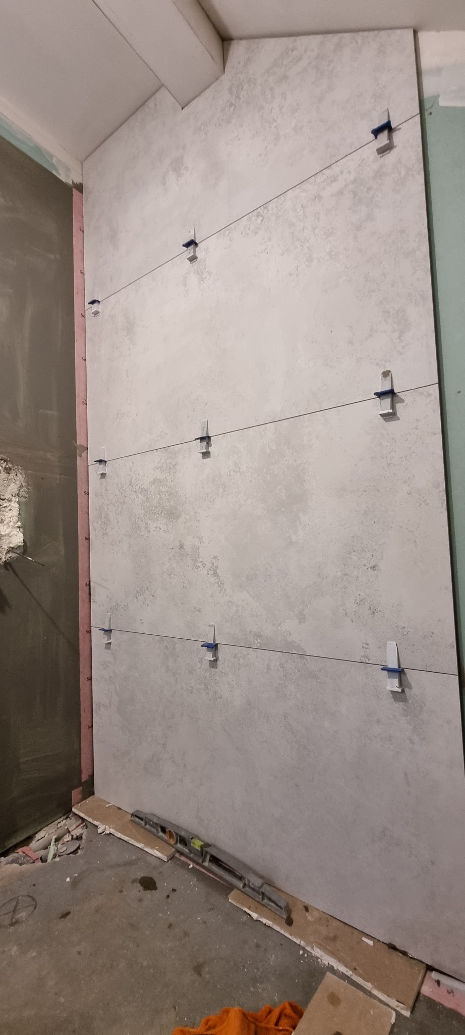 Układanie wielkoformatowych płytek imitujących beton na ścianie z użyciem poziomicy laserowej i klipsów poziomujących; widoczna warstwa izolacji po lewej stronie.