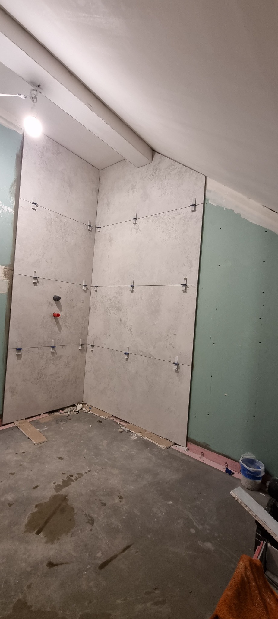 Montaż szarych płytek wielkoformatowych w rogu łazienki, widoczne krzyżyki dystansowe i poziomowanie laserowe, surowy beton na podłodze i ściana z płyt gipsowo-kartonowych w tle.