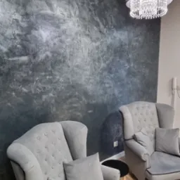 Dwa szare fotele typu uszak z podnóżkiem i poduszkami, ustawione na tle ściany z dekoracyjną szarą fakturą i oświetlone kryształowym żyrandolem.