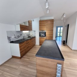 RAFAŁ KAŹMIERSKI - Nowoczesna kuchnia z drewnianymi frontami szafek, czarnym blatem i marmurowym backsplashiem, oświetlona reflektorami, z wyspą kuchenną z gniazdkami i psem w tle.