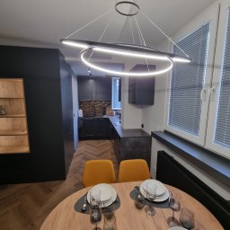 RAFAŁ KAŹMIERSKI - Nowoczesna kuchnia z czarnymi szafkami, drewnianym blatem i podłogą w jodełkę, oświetlona designerską lampą wiszącą nad okrągłym stołem z nakryciem dla czterech osób.
