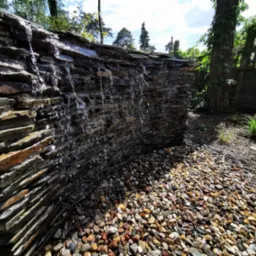 Kamienna kaskada wodna w ogrodzie, woda spływa po ułożonych warstwowo płytach skalnych, dół wysypany kolorowymi kamykami, w tle zieleń drzew i krzewów.