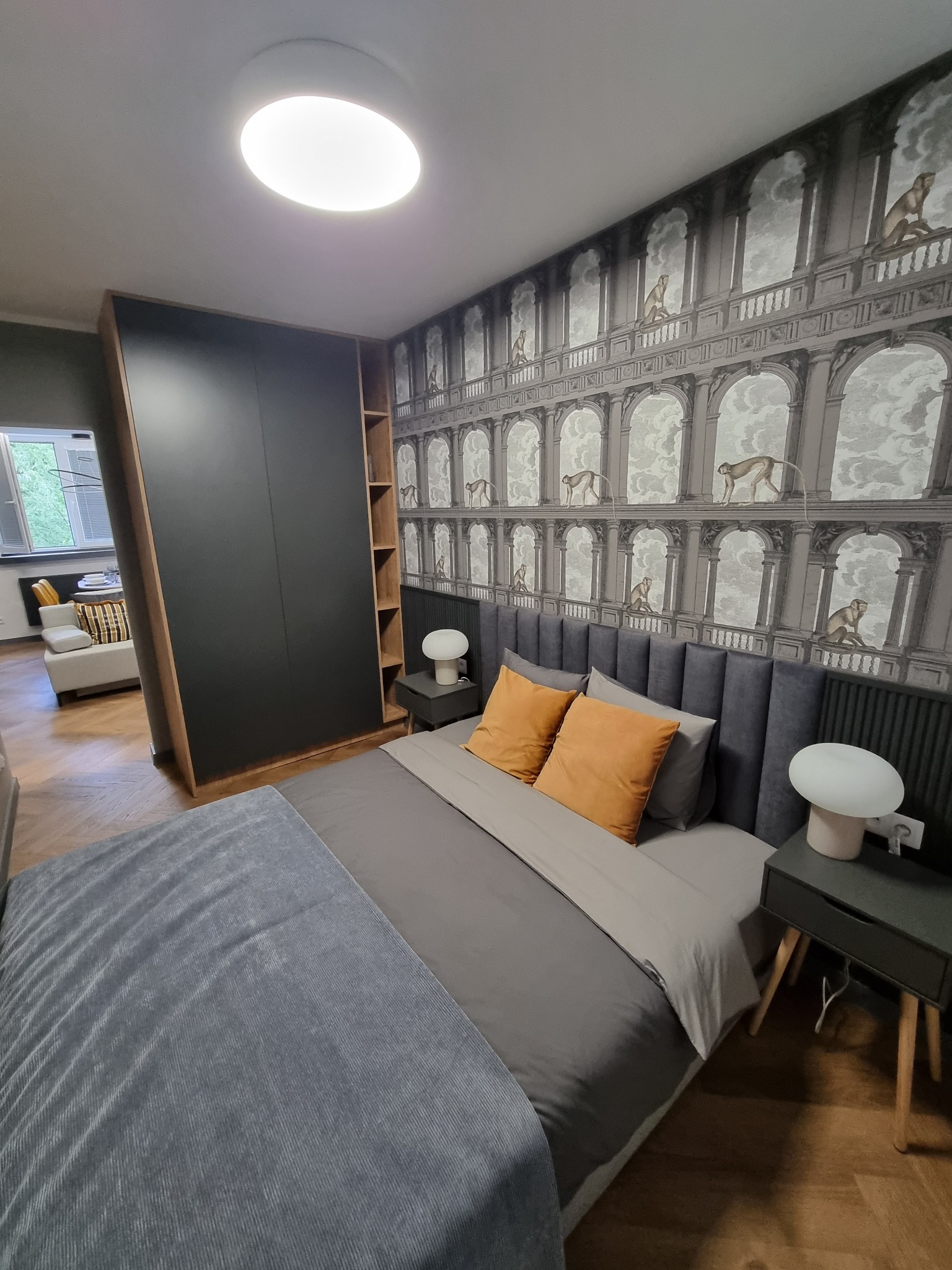 Sypialnia z łóżkiem z szarą tapicerowaną ramą, pomarańczowymi poduszkami i szafkami nocnymi z lampkami, na ścianie tapeta z motywem architektury i małp.