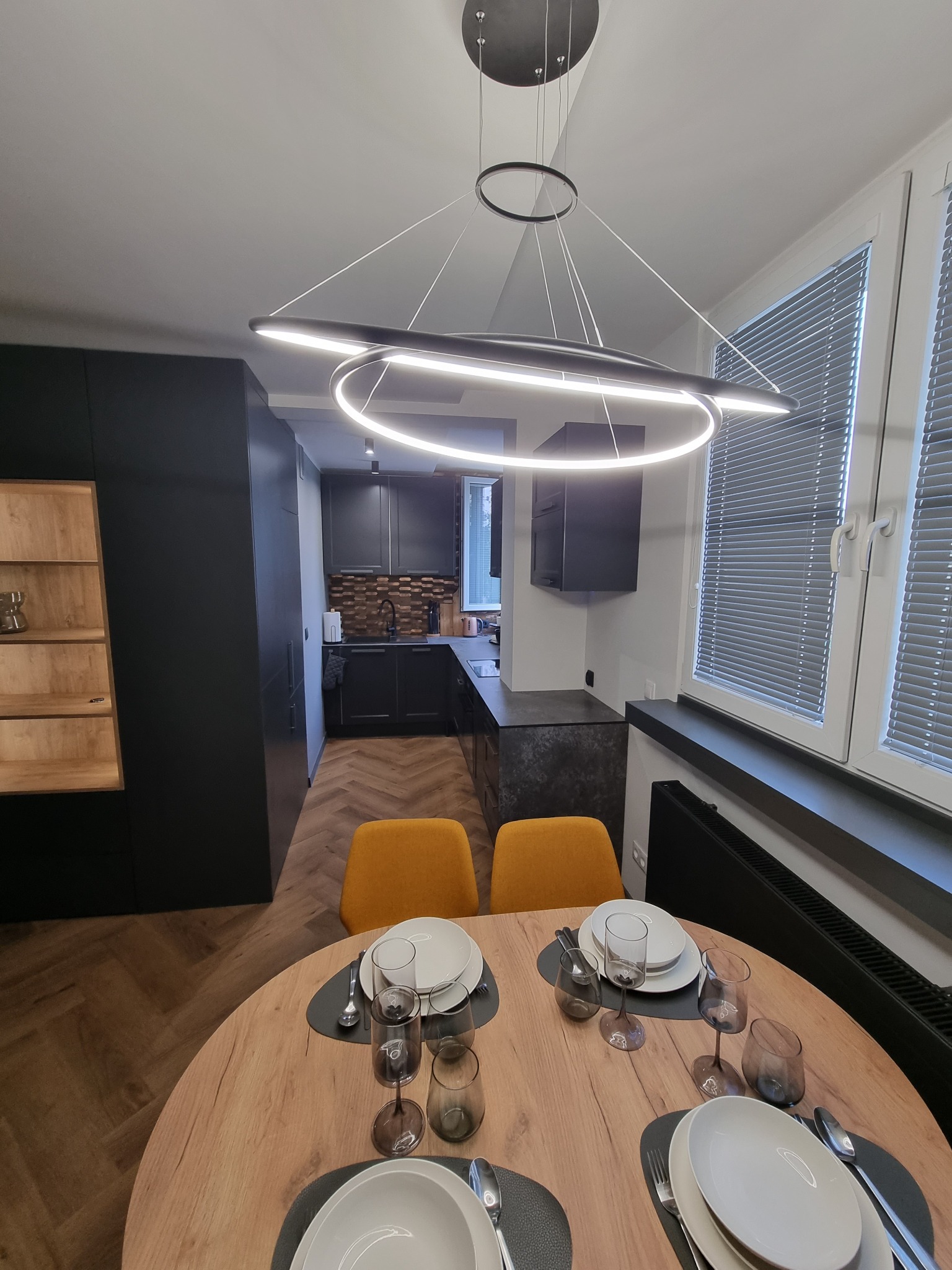 Nowoczesna kuchnia z czarnymi szafkami, drewnianym blatem i podłogą w jodełkę, oświetlona designerską lampą wiszącą nad okrągłym stołem z nakryciem dla czterech osób.