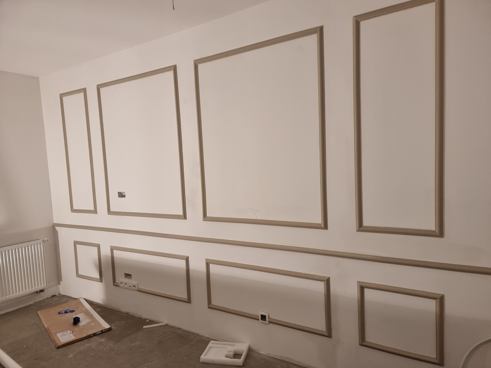Ściana z zamontowanymi sztukateriami w formie paneli z listew, widoczne gniazdka elektryczne, kaloryfer i karton na podłodze w trakcie prac wykończeniowych.
