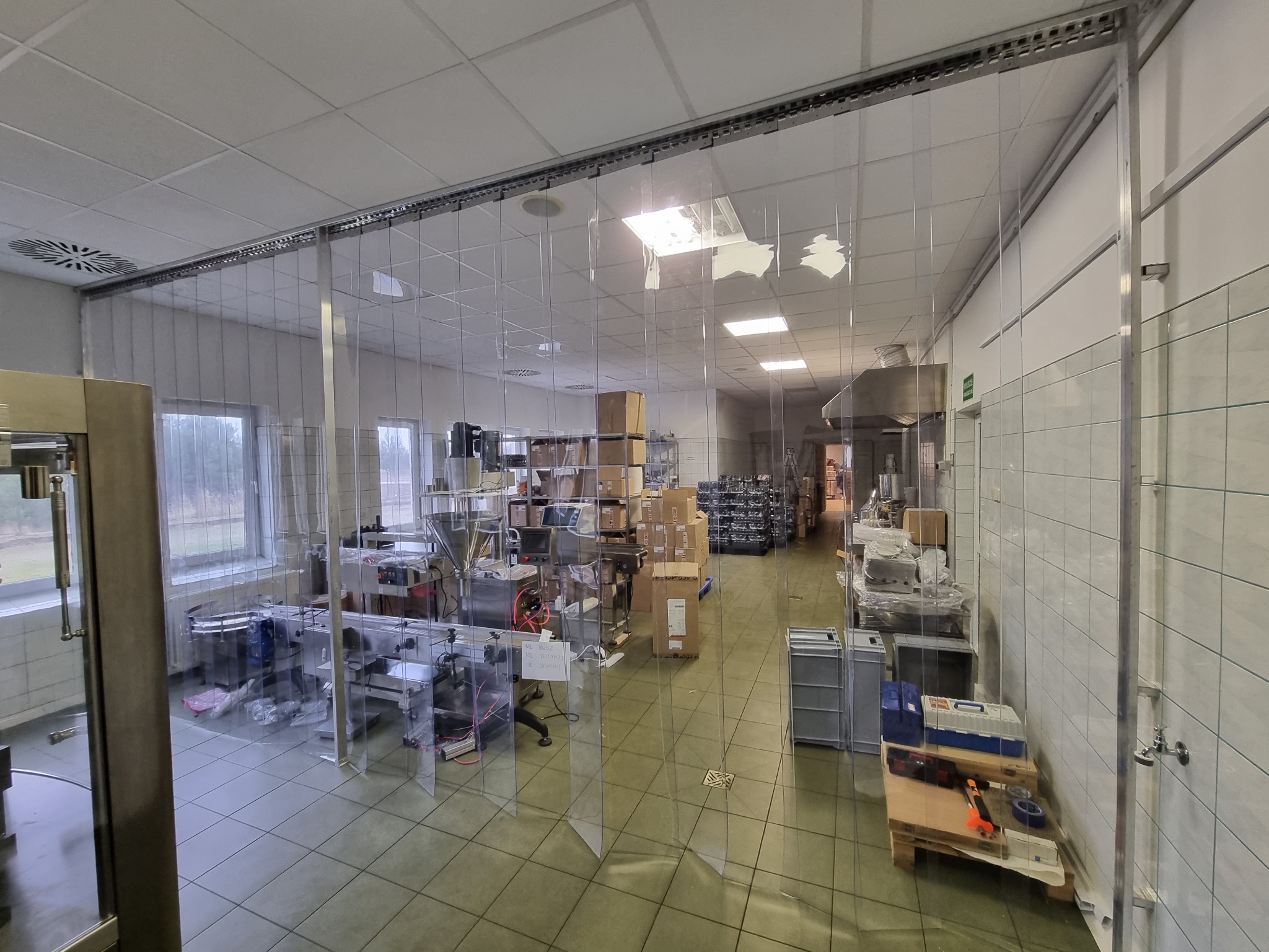 Widok wnętrza hali produkcyjnej oddzielonej kurtyną paskową z PCV, widoczne maszyny, urządzenia i materiały w kartonach.