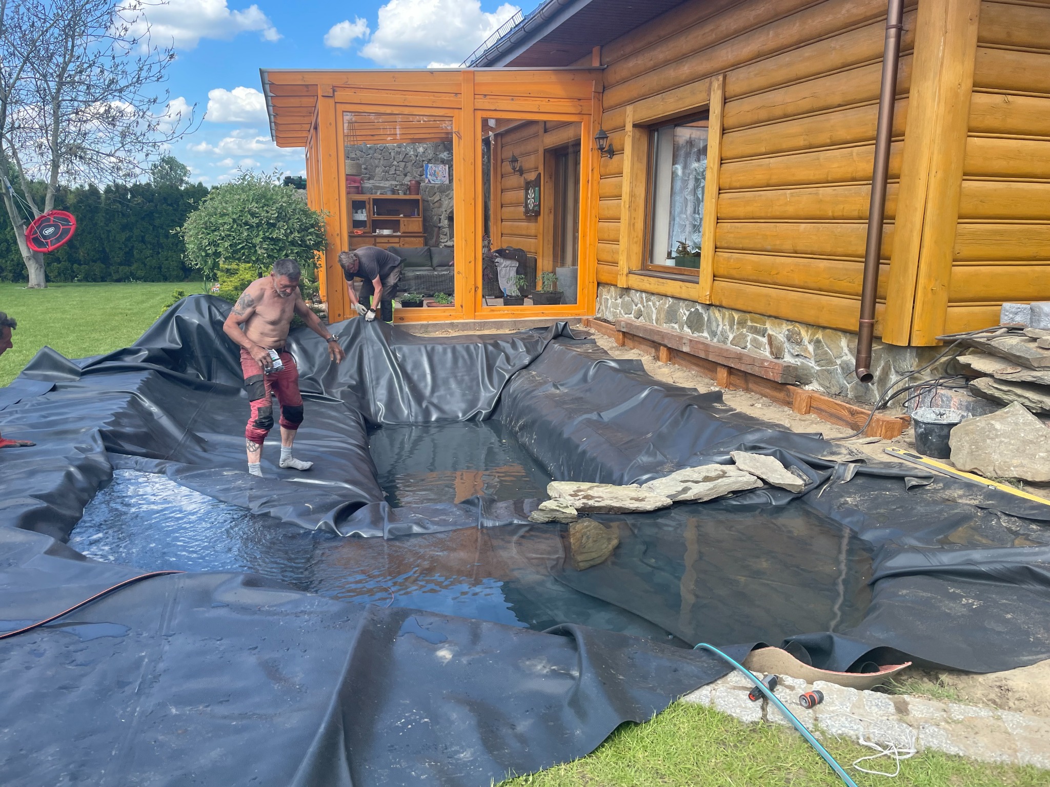 Proces tworzenia oczka wodnego z czarną folią w ogrodzie przy drewnianym domu, dwóch mężczyzn w trakcie pracy, jeden w czerwonych spodniach stoi w wodzie, drugi w szarych rękawicach pracuje na brzegu.