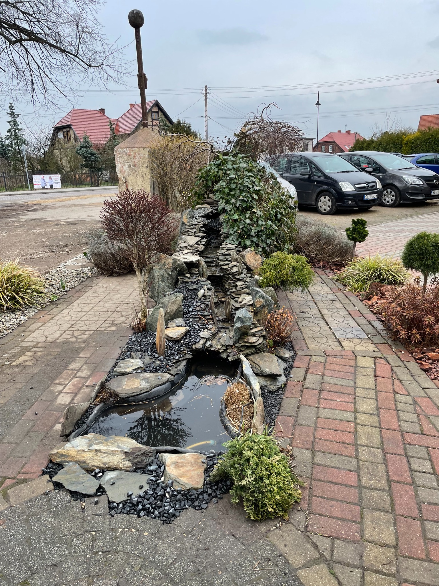 Ogród z kaskadą wodną wykonaną z kamieni, otoczony różnorodną roślinnością ozdobną i brukowaną ścieżką, z widocznymi samochodami i budynkami w tle