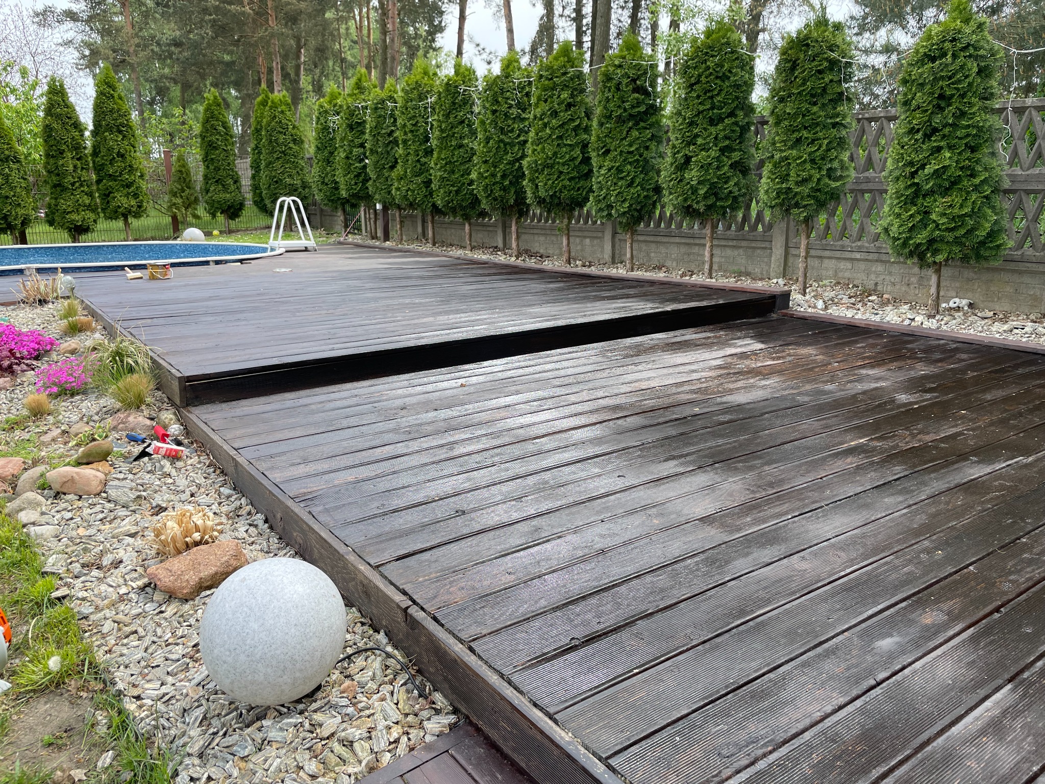 Ciemnobrązowy, drewniany taras przylegający do basenu, otoczony tujami i kamieniami ozdobnymi, z widoczną lampą ogrodową w kształcie kuli.