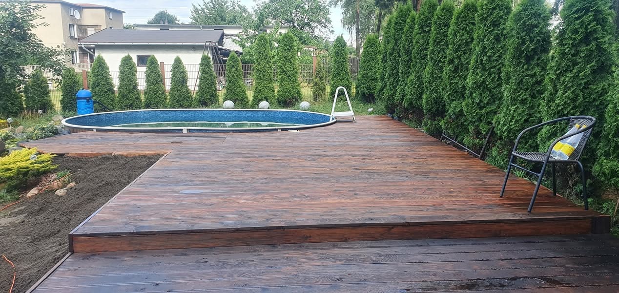 Brązowy taras drewniany z widocznym basenem ogrodowym, rzędem tuj i krzesłem z butelką wody mineralnej.