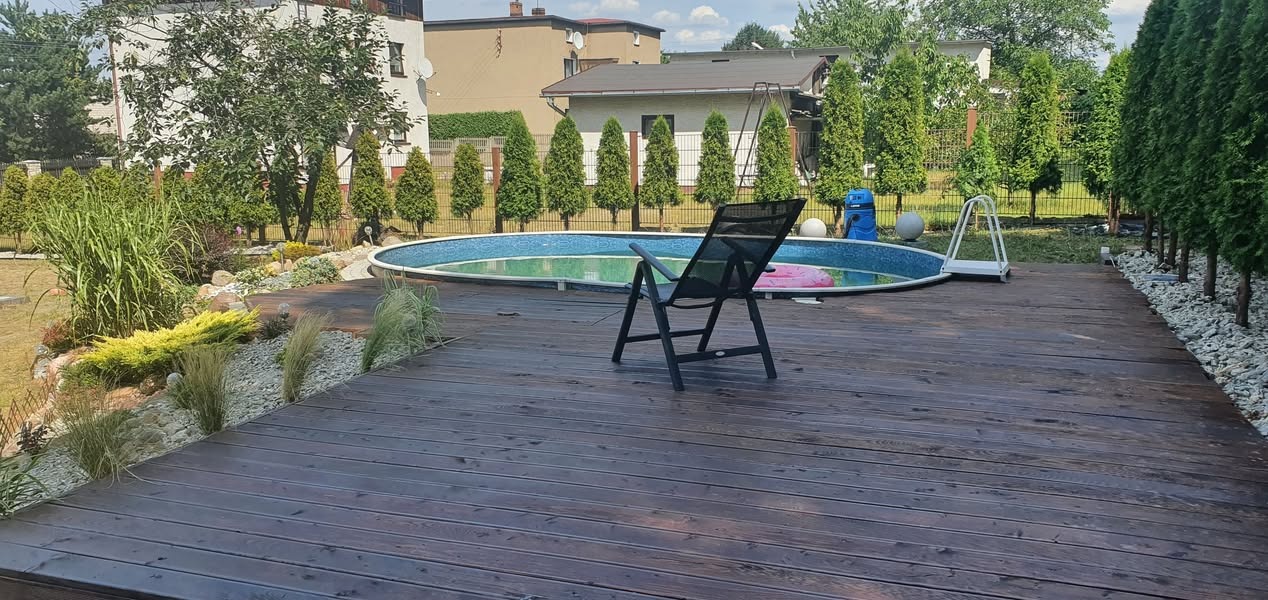 Ciemnobrązowy taras drewniany z widokiem na okrągły basen ogrodowy, otoczony kamieniami i tujami, z czarnym składanym krzesłem na pierwszym planie.