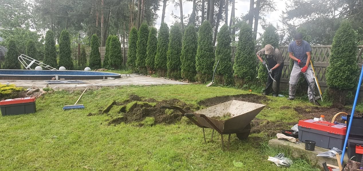 Dwaj mężczyźni kopią ziemię w ogrodzie obok basenu, taczka z trawą, narzędzia w skrzynkach, rząd tuj w tle.