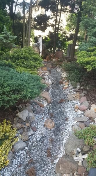 Ogród z kamiennym korytem wypełnionym drobnym żwirem, otoczonym roślinnością i kamieniami, z widocznymi elementami dekoracyjnymi w tle.