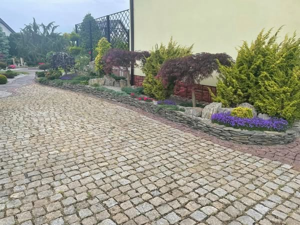 Wyłożona kostką brukową ścieżka prowadzi przez zadbany ogród z ozdobnymi kamieniami, karłowatymi drzewami i kolorowymi kwiatami wzdłuż ściany budynku.