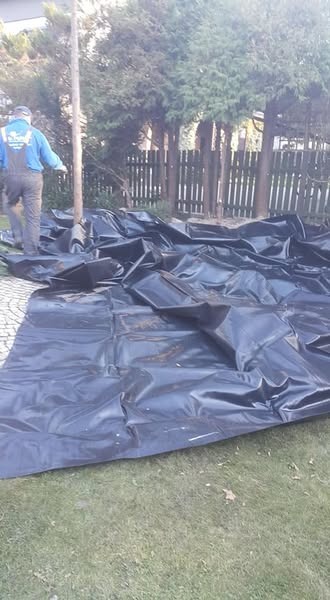 Czarna folia rozłożona na trawie, prawdopodobnie pod oczko wodne, z widocznym pracownikiem w kombinezonie na tle drewnianego płotu i drzew.