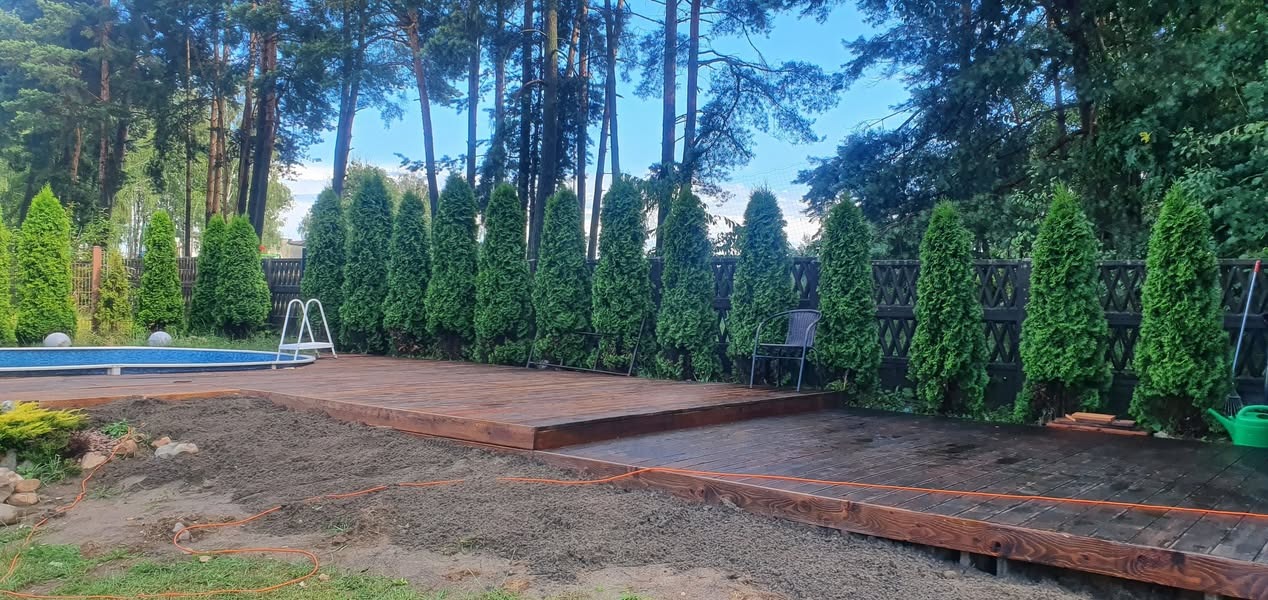 Drewniany taras z desek przy basenie i szpalerze tuj, widoczny fragment przygotowanego terenu pod nasadzenia.