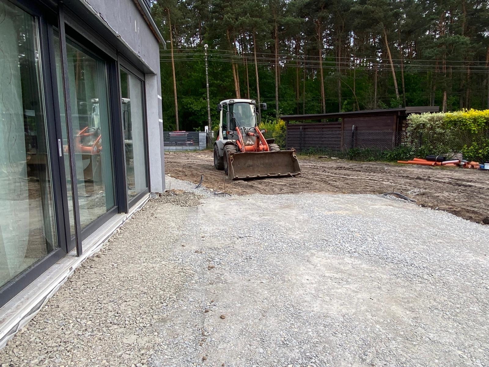 Przygotowanie podłoża pod kostkę brukową: miniładowarka rozplantowuje żwir obok nowoczesnego domu z dużymi przesuwnymi oknami, w tle widoczne rury kanalizacyjne i drzewa.