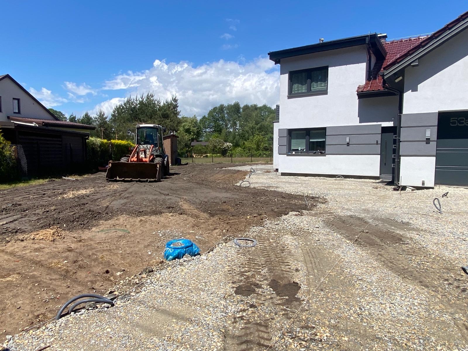 Przygotowanie terenu pod budowę lub ogród: miniładowarka wyrównuje ziemię, widoczne kable i worki z materiałami budowlanymi, w tle dom z szarą elewacją.