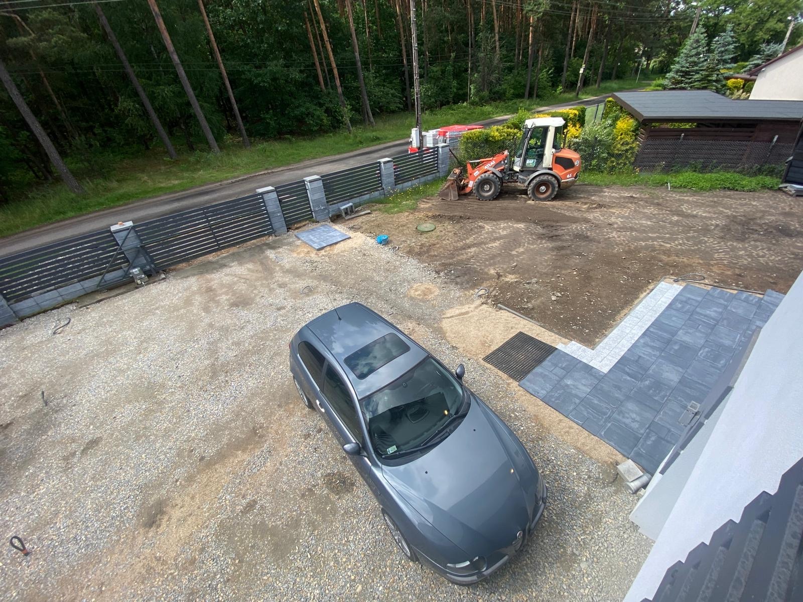 Widok z góry na teren posesji w trakcie prac ziemnych, z zaparkowanym szarym samochodem, fragmentem ułożonej kostki brukowej oraz koparką przygotowującą podłoże, otoczoną zielenią i lasem w tle.