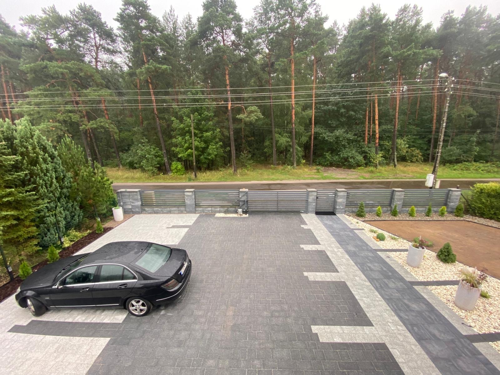 Nowoczesny podjazd z szarej kostki brukowej ułożonej we wzór, z zaparkowanym czarnym samochodem, ogrodzeniem z szarych paneli i kolumn oraz widokiem na las w tle.