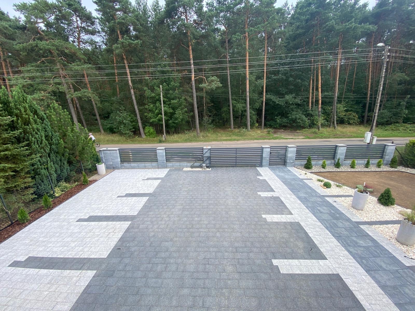 Ułożona kostka brukowa w odcieniach szarości i bieli, tworząca geometryczny wzór na podjeździe, z widocznym fragmentem ogrodzenia panelowego i zielenią drzew w tle.