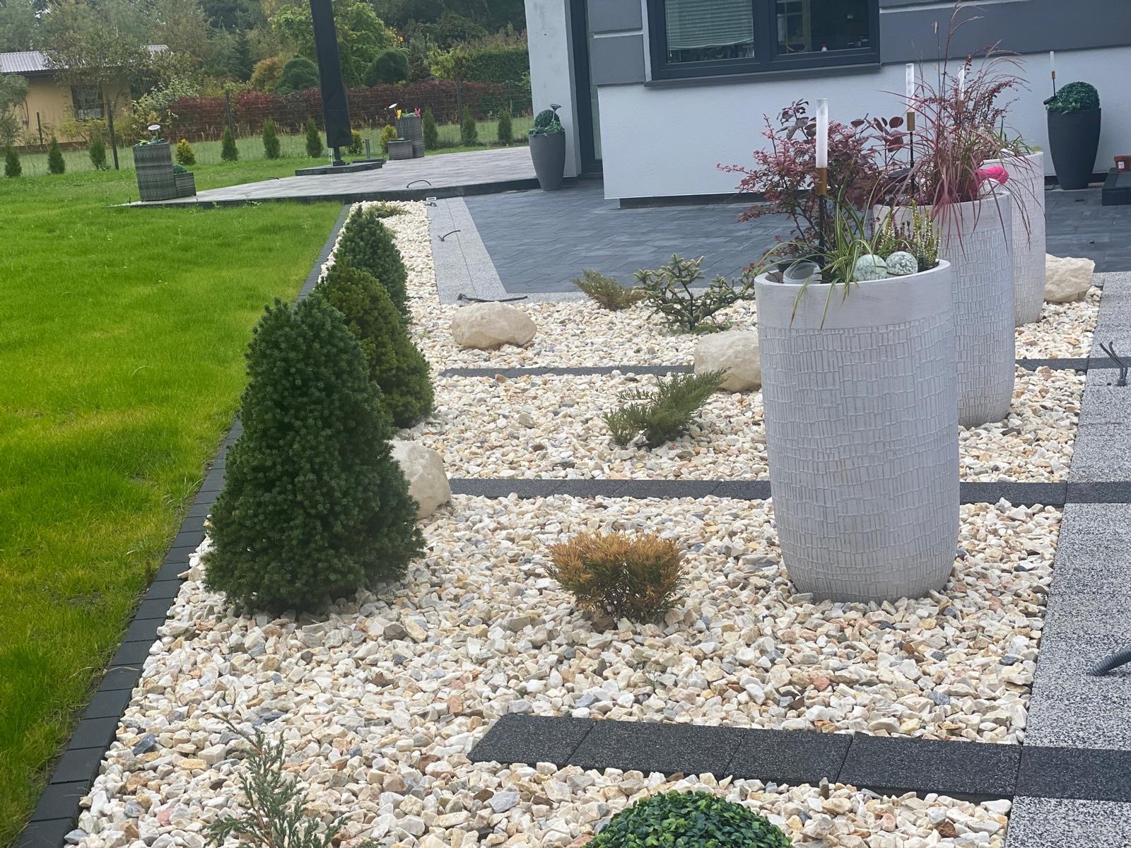 Ogród z białym żwirem, ozdobnymi kamieniami, niskimi iglakami, donicami z roślinami i ścieżką z ciemnych płyt, widok z perspektywy niskiej.