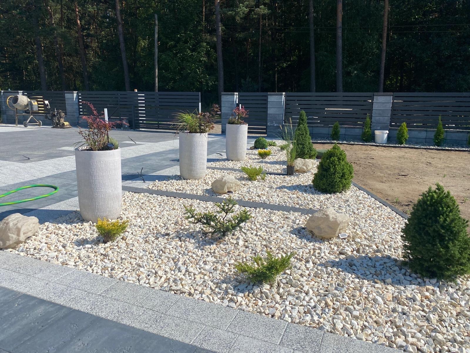 Nowoczesny ogród z białym żwirem, roślinami w betonowych donicach i niskimi iglakami, obrzeża z szarej kostki, w tle ciemne ogrodzenie panelowe i drzewa.